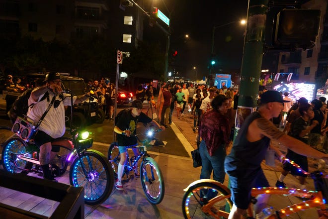 Colorfully lit bikes go by as people fill the street for the First Friday art walk on Roosevelt Row on Aug. 4, 2023, in downtown Phoenix.
